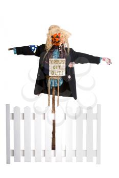pretty scarecrow in a jacket with patches and a straw hat with a sign that is sure to scare away the crows is behind the fence. Isolated on white