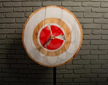 Rough wooden target with a single arrow caught right in the center of a red circle against a gray brick wall