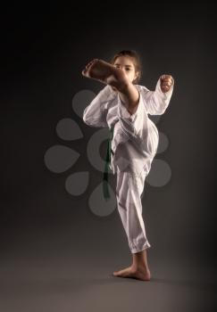 A little girl in a traditional white kimono for karate and a green belt performs training exercises against a dark background.