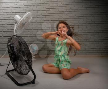 A little girl cools herself from the heat with the help of three different fans at once sitting on the floor of the room