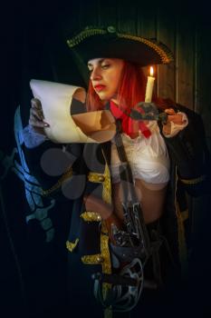 Young Attractive armed girl pirate captain examines a map by candlelight against the background of the flag Jolly Roger