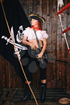 Young attractive armed girl pirate captain looks into the distance on the background of the flag Jolly Roger