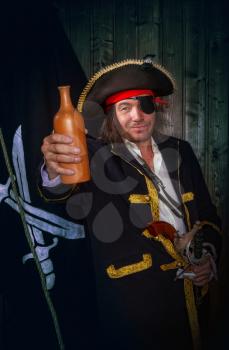 Adult pirate captain in a traditional costume and with weapons drinks rum from a clay bottle against the background of a jolly roger