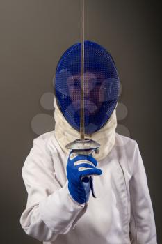 little boy fencer armed with a rapier in white sports equipment and with a protective helmet on a dark background