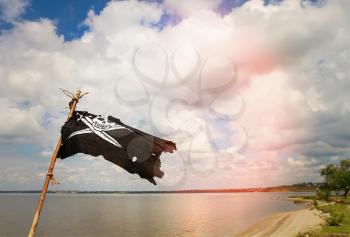Black torn pirate flag Jolly Roger with crossed sabers waving on a stick against the background of the river and cloudy sky