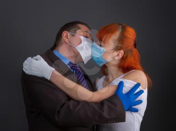 A man in a suit and a girl kiss on a dark background through protective medical masks and gloves.