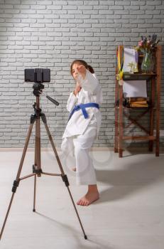A little girl in a white kimono with a blue belt takes karate lessons via the Internet while at home on self-isolation during quarantine