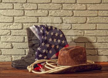 traditional wide-brimmed cowboy hat, rough lasso boots and a striped USA flag on a brick background