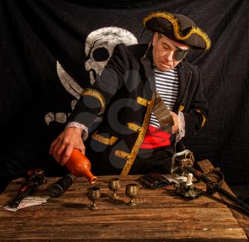 An adult pirate captain drinks alcohol from a clay bottle in a glass against the background of a black flag Jolly Roger