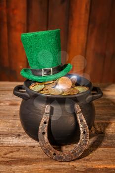 Treasure in a black pot and a green leprechaun hat on a wooden table next to a horseshoe symbol of good luck