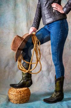 Female legs in leather cowboy boots and blue jeans with wide-brimmed hat and lasso on grunge background