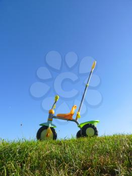 Childhood toy tricycle standing on green grass with blue sky background.