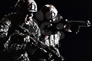 Half length low angle studio shot of pair two special forces soldiers in field uniforms with weapons attacking shouting, portrait on black background