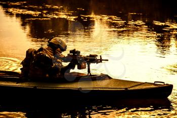 Special forces backlit machine gunner in army kayak. Boat moving calmly across the river, diversionary mission, sunset dusk