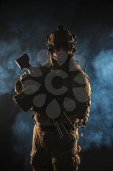 Army soldier in Combat Uniforms with machine gun, plate carrier and combat helmet are on. Studio contour silhouette shot, backlight, dark glowing smoke background