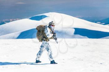 Army serviceman in winter camo somewhere in the Arctic. He wears chest rig and huge backpack, suffers from the extreme cold and strong wind, but endures while mission continues. Blue sky background