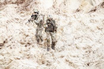 Two infantry soldiers in combat camo uniform, with tactical ammunition, carefully walking and aiming with assault rifles in unknown desert area. Military reconnaissance, scouting on enemy territory