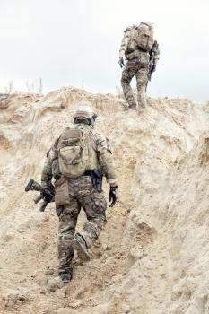Two modern infantry, special operations forces soldiers, military intelligence specialists armed with service rifles in protective camouflage uniform with backpack on backs climbing on sand dune