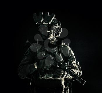 Army special operations forces soldier in mask and combat uniform, helmet equipped night-vision device, armed submachine gun with silencer, looking aside, low key studio portrait on black, copyspace