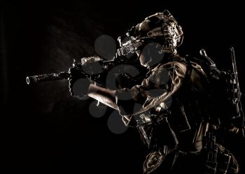 Army special operations soldier, commando fighter in full tactical ammunition, helmet with radio headset and night vision device, aiming short barrel assault rifle in darkness, low key studio shoot