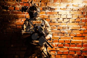 Special operations forces soldier, counter terrorism assault team fighter with night vision device on helmet and service rifle, low key indoor shot brick wall