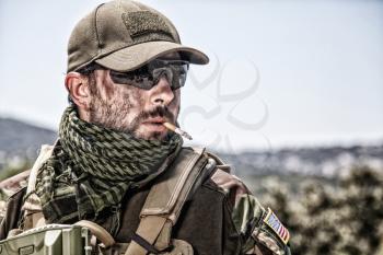 Shoulder portrait of commando fighter, professional mercenary or special forces shooter with dirty face, wearing shemagh scarf, ballistic glasses and cap, carrying service rifle and smoking cigarette