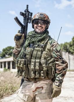 Army soldier, special forces infantryman, Navy SEALs fighter in camouflage uniform, combat helmet and plate carrier, armed submachine gun. Half-length portrait of happy smiling commando shooter