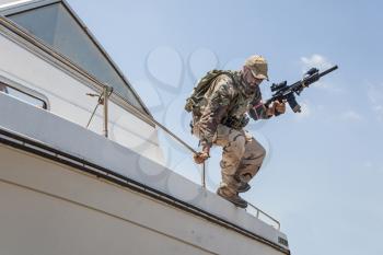 Army commando, special forces soldier equipped with body armour and battle helmet with radio headset, screaming and yelling while jumping from speed boat deck, landing on shore with assault rifle