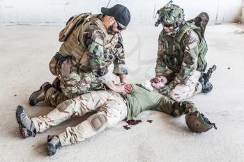 Soldiers trying to stop bleeding at wounded comrade who lying on floor, suffering and screaming in paine. Commando fighter pressing with hands on bloody wound at friends stomach, giving emergency care