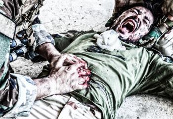 Wounded soldier, commando fighter lying on floor, screaming in pain while comrade pressing with hands on wound in his stomach, trying stop bleeding and save life. Tactical casualty care on battlefield