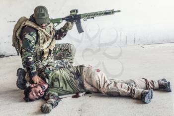 Army soldier, special forces fighter in ammunition, armed assault rifle, kneeling and placing two fingers on comrades neck to feel pulse. Shooter checking heartbeat of wounded in gunfight friend