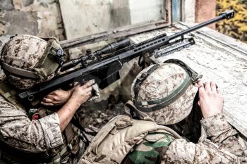 Marines sniper team armed with large caliber, anti-materiel sniper rifle hiding in ruined urban building, shooting enemy targets on range from shelter, sitting in ambush. Military firefight in city
