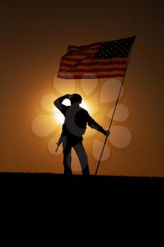 Silhouette of US army soldier, Marines Corps fighter or special forces rifleman in helmet, armed rifle standing on hill with waving on wind national flag, looking far away on background of sunset sky