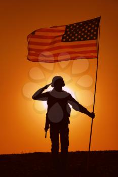 Silhouette of USA armed forces soldier, army infantryman or Marine Corps fighter saluting while standing with waving national flag on sunset background. Military victory and glory, fallen remembrance