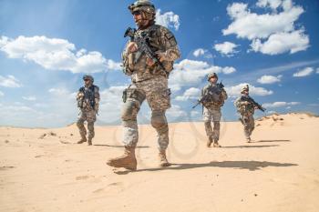 Team of United states airborne infantry men with weapons moving patrolling desert. Sand and blue sky on background of squad, sunlight, low angle view