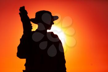 Silhouette of army special forces soldier in boonie and ballistic goggles, standing with bullpup submachine gun on background of sunset sky. Commando fighter, elite forces rifleman patrolling on dawn