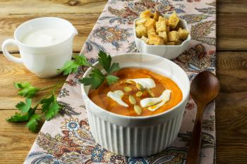 Autumnal spicy pumpkin squash cream soup with cream, parsley and pumpkin seeds and in the white bowl, cream in a creamer and croutons in a serving dish on the napkin on rustic wooden background
