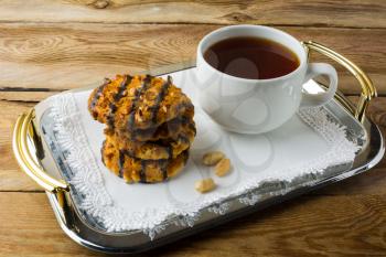 Chocolate icing cookies with peanuts and cup of tea on a metal serving tray with handles covered with a white lace napkin. Breakfast biscuits and tea.