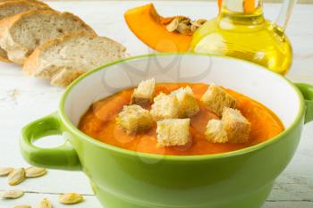 Creamy pumpkin squash vegetable soup with croutons in a green bowl and bread on white wooden background