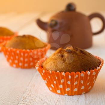 Muffins in orange paper cupcake holder with white polka dots and brown ceramic teapot on a white wooden background, selective focus, square 
