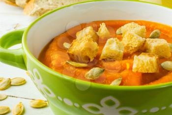Pumpkin squash vegetable soup with croutons and pumpkin seeds in a green bowl on white wooden background, close up