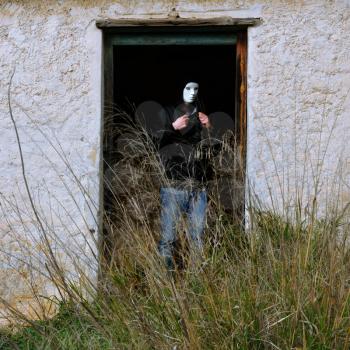Man with white mask by broken door of abandoned house and overgrown plants.