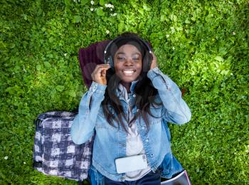 Top view of smiling african woman lying on green grass listening to music with headphones.